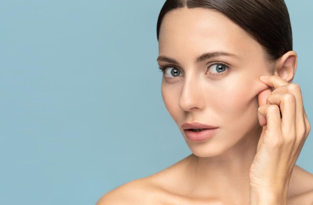 A person with clear skin gently pinching the skin near her cheekbone, demonstrating facial skin tightness against a light blue background.