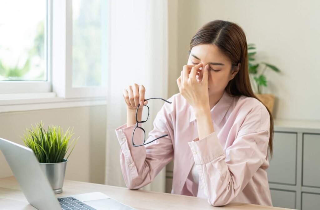 A person sits at a desk holding a pair of glasses and pinching the bridge of their nose, suggesting eye strain or discomfort while working on a laptop.