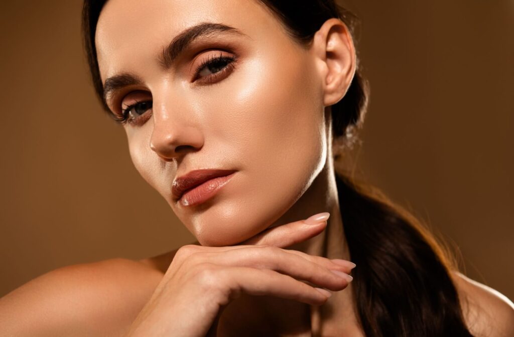 A close-up portrait of a woman with smooth, glowing skin, resting her chin on her hand against a warm, neutral background