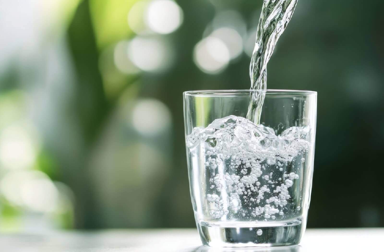 A clear glass being filled with fresh water, bubbles forming as it pours, set against a soft green, out-of-focus background that suggests freshness and hydration.