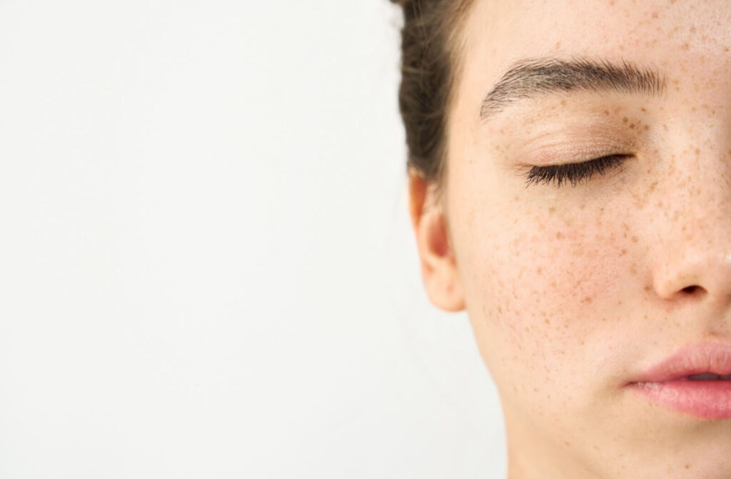 A close-up, cropped view of a woman's face with her eyes closed, showing natural freckles on her cheek and nose area.