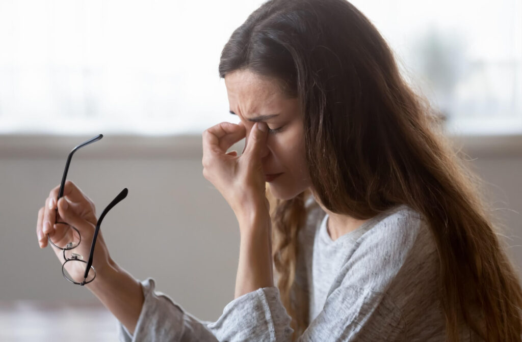 A side profile of an individual holding their eyeglasses while pinching the bridge of their nose and closing their eyes in discomfort.