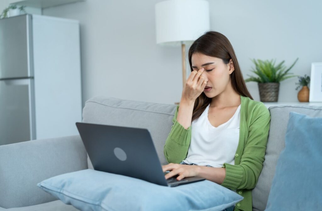 An individual in a green cardigan sitting on a sofa with a laptop, pinching the bridge of their nose with closed eyes.