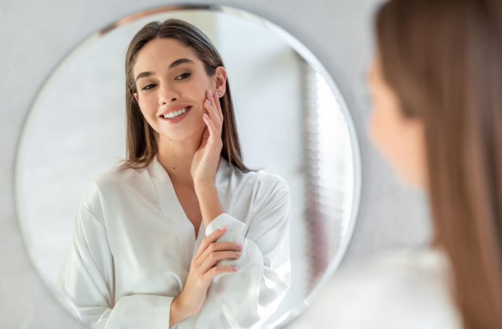 A happy woman in a white silk robe looking at her reflection in a circular mirror and touching her firm, youthful-looking cheek after a facial treatment.